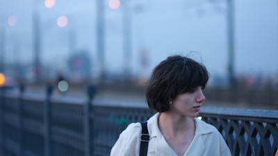 A person with short dark hair stands next to a metal railing. The background is blurred, featuring soft lights and hints of distant structures.