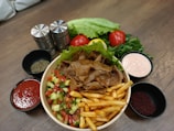 A wooden table featuring a bowl filled with meat slices, French fries, and a mixed salad of diced cucumbers, tomatoes, and herbs. Surrounding the bowl are small bowls containing red sauce, pink sauce, and various spices. Fresh ingredients like whole tomatoes, lettuce, and leafy greens are placed at the back, along with salt and pepper shakers.
