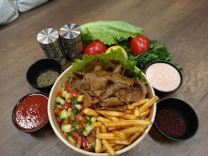 A wooden table featuring a bowl filled with meat slices, French fries, and a mixed salad of diced cucumbers, tomatoes, and herbs. Surrounding the bowl are small bowls containing red sauce, pink sauce, and various spices. Fresh ingredients like whole tomatoes, lettuce, and leafy greens are placed at the back, along with salt and pepper shakers.