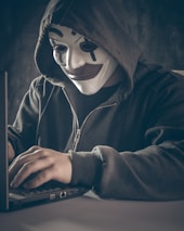 A person wearing a symbolic mask with a dark hoodie is using a laptop. The scene is dimly lit, creating a sense of mystery and anonymity.