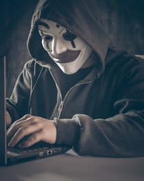 A person wearing a symbolic mask with a dark hoodie is using a laptop. The scene is dimly lit, creating a sense of mystery and anonymity.