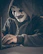 A person wearing a symbolic mask with a dark hoodie is using a laptop. The scene is dimly lit, creating a sense of mystery and anonymity.
