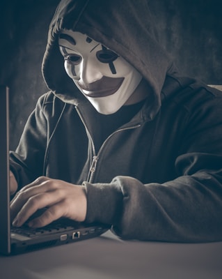 A person wearing a symbolic mask with a dark hoodie is using a laptop. The scene is dimly lit, creating a sense of mystery and anonymity.