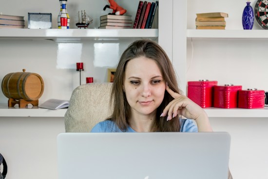 A woman is sitting in a comfortable chair in front of a laptop, appearing focused and engaged. Books and decorative items, including a small barrel and various colored containers, are neatly arranged on the shelves behind her.