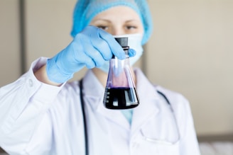 A person wearing a lab coat, mask, and blue gloves holds up an Erlenmeyer flask containing a dark liquid for examination. They appear to be in a laboratory setting.