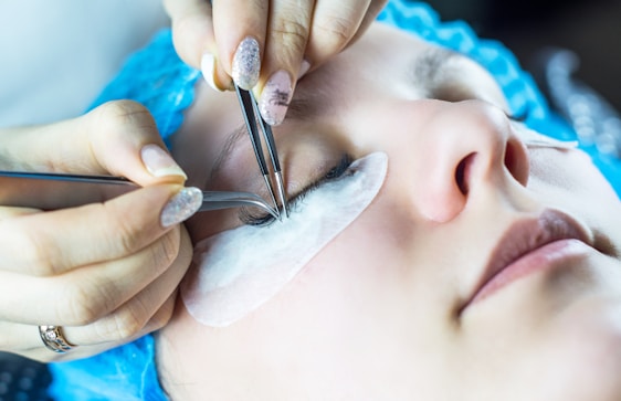 A person is receiving an eyelash extension treatment. They are lying down with closed eyes, and two hands are applying the extensions using tweezers. Eye pads are placed underneath the eyes for protection.