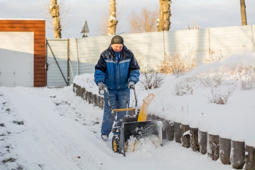 A person operates a snowblower on a snow-covered path next to a wooden fence and barren shrubs under a cloudy sky. The person is dressed in a blue winter jacket and a hat, and the machine is actively clearing the snow.