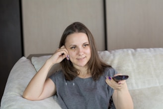 A young woman with brown hair is sitting on a light-colored couch. She holds a remote control in her right hand and rests her head on her left hand. The room features neutral-toned walls and a cozy atmosphere.