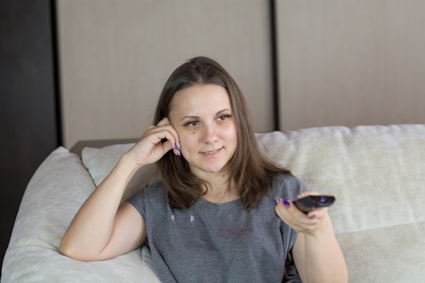 A young woman with brown hair is sitting on a light-colored couch. She holds a remote control in her right hand and rests her head on her left hand. The room features neutral-toned walls and a cozy atmosphere.
