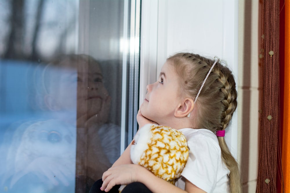 a little girl looking out a window with a stuffed animal