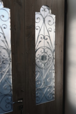 Two wooden doors with frosted glass panels featuring intricate wrought iron patterns. The sunlight casts clear shadows onto the panels, highlighting the ornamental scrollwork. A metal door handle is affixed to the door on the left, with visible grain textures in the wood.