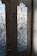 Two wooden doors with frosted glass panels featuring intricate wrought iron patterns. The sunlight casts clear shadows onto the panels, highlighting the ornamental scrollwork. A metal door handle is affixed to the door on the left, with visible grain textures in the wood.