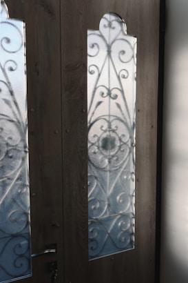 Two wooden doors with frosted glass panels featuring intricate wrought iron patterns. The sunlight casts clear shadows onto the panels, highlighting the ornamental scrollwork. A metal door handle is affixed to the door on the left, with visible grain textures in the wood.