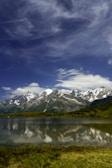 Snow-capped mountains reflecting in a calm alpine lake under a bright blue sky.