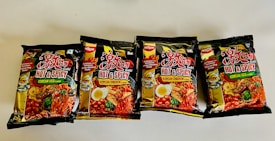 Four packs of instant noodles are displayed on a white surface. The packaging is black with vibrant images and text. The labels indicate two different flavors: 'Hot & Spicy Korean Veg' and 'Hot & Spicy Korean Chicken'. The packaging prominently features images of noodles, vegetables, and spices, highlighting the spicy nature of the products. There are small illustrations of a cartoon character on the packaging as well.
