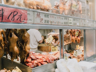 A variety of meats, including roasted ducks and pork, are displayed in a shop window. Price tags written in both Chinese characters and numbers hang from the meat. The display is vibrant with cooked and raw meats, and some parts of the interior of the shop are visible.