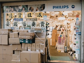 A store entrance with a prominent display of lighting fixtures. The store has a signboard with Asian characters and the word 'PHILIPS' along with the LED symbol. Numerous cardboard boxes are stacked outside the entrance, possibly containing merchandise. Inside, there are several lighting products on display, and two people, a man and a woman, are seen working.