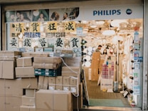 A store entrance with a prominent display of lighting fixtures. The store has a signboard with Asian characters and the word 'PHILIPS' along with the LED symbol. Numerous cardboard boxes are stacked outside the entrance, possibly containing merchandise. Inside, there are several lighting products on display, and two people, a man and a woman, are seen working.