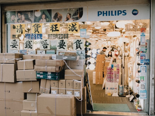 A store entrance with a prominent display of lighting fixtures. The store has a signboard with Asian characters and the word 'PHILIPS' along with the LED symbol. Numerous cardboard boxes are stacked outside the entrance, possibly containing merchandise. Inside, there are several lighting products on display, and two people, a man and a woman, are seen working.