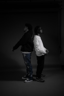 Two individuals stand back to back in a dimly lit setting, each with their hands clasped in front. One wears a dark hoodie and jeans, while the other is in a white jacket and dark pants. The overall atmosphere is minimalistic and moody.