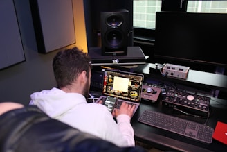 A person seated at a desk is working on a laptop with various audio equipment surrounding them. The setup includes large studio speakers, an external audio interface, and a keyboard. The laptop screen displays a software application with multiple windows open. The room has a professional and tech-focused atmosphere.