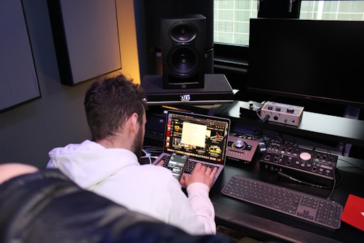 A person seated at a desk is working on a laptop with various audio equipment surrounding them. The setup includes large studio speakers, an external audio interface, and a keyboard. The laptop screen displays a software application with multiple windows open. The room has a professional and tech-focused atmosphere.