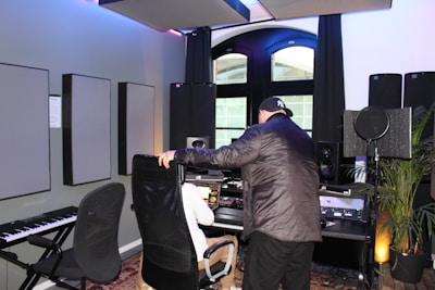 Two people are present in a well-equipped music studio. One person is seated at a computer desk with various audio equipment, including speakers and a microphone. The other person is leaning over the seated individual while interacting with them. There is also a keyboard and acoustic panels on the walls. The room has modern decor and large windows with natural light coming through.