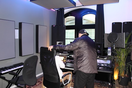 Two people are present in a well-equipped music studio. One person is seated at a computer desk with various audio equipment, including speakers and a microphone. The other person is leaning over the seated individual while interacting with them. There is also a keyboard and acoustic panels on the walls. The room has modern decor and large windows with natural light coming through.