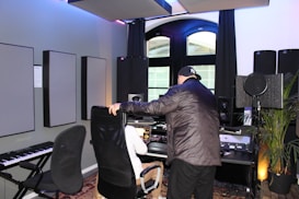 Two people are present in a well-equipped music studio. One person is seated at a computer desk with various audio equipment, including speakers and a microphone. The other person is leaning over the seated individual while interacting with them. There is also a keyboard and acoustic panels on the walls. The room has modern decor and large windows with natural light coming through.
