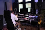 A person wearing a white jacket is seated in a black chair and is working on a digital audio workstation in a music studio. The studio contains multiple speakers, a large monitor, various sound mixing equipment, and headphones. There is also a microphone setup with a pop filter, and plants in the background add a touch of greenery.