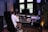A person wearing a white jacket is seated in a black chair and is working on a digital audio workstation in a music studio. The studio contains multiple speakers, a large monitor, various sound mixing equipment, and headphones. There is also a microphone setup with a pop filter, and plants in the background add a touch of greenery.