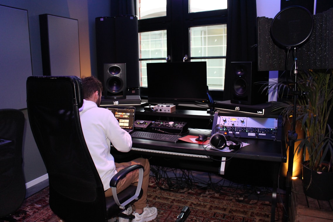 A person wearing a white jacket is seated in a black chair and is working on a digital audio workstation in a music studio. The studio contains multiple speakers, a large monitor, various sound mixing equipment, and headphones. There is also a microphone setup with a pop filter, and plants in the background add a touch of greenery.