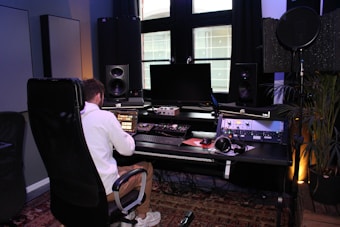 A person wearing a white jacket is seated in a black chair and is working on a digital audio workstation in a music studio. The studio contains multiple speakers, a large monitor, various sound mixing equipment, and headphones. There is also a microphone setup with a pop filter, and plants in the background add a touch of greenery.