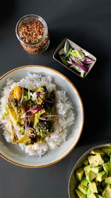 A bowl filled with fluffy white rice topped with roasted vegetables such as broccoli and onions, garnished with chili flakes. Nearby, a small rectangular dish holds sliced green and purple onions, while a jar contains red chili flakes. A separate bowl holds diced avocado pieces.