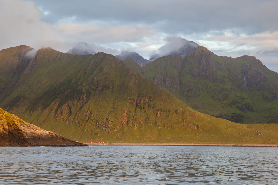 The image depicts a serene mountainous landscape with a large body of water at its base, possibly a lake or river. The mountains are covered in lush green vegetation and have snow-capped peaks that add to their majestic appearance. The sky above is filled with clouds, casting a soft light over the scene. In the foreground, there's a small island visible on the water, adding depth to the image.