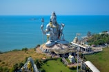 A massive statue of a deity is prominently displayed on a coastal landscape, surrounded by lush greenery and overlooking the sea. The figure is seated with a serene expression, holding a trident in one hand. The area is bustling with visitors and includes several smaller structures and pathways.