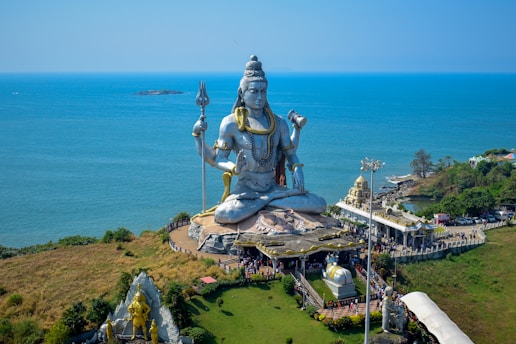 A massive statue of a deity is prominently displayed on a coastal landscape, surrounded by lush greenery and overlooking the sea. The figure is seated with a serene expression, holding a trident in one hand. The area is bustling with visitors and includes several smaller structures and pathways.
