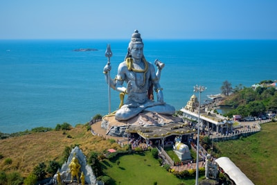 A massive statue of a deity is prominently displayed on a coastal landscape, surrounded by lush greenery and overlooking the sea. The figure is seated with a serene expression, holding a trident in one hand. The area is bustling with visitors and includes several smaller structures and pathways.