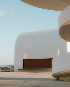 A modern architectural scene featuring smooth, curving white structures against a clear blue sky. The building has a minimalist design with clean lines and an open space in the foreground. A large red rectangular element provides contrast to the otherwise monochromatic setting.
