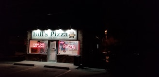 A small brick storefront is illuminated at night by overhead lights. The sign reads 'Bill's Pizza EST. 1957' with a pizza graphic, and there are lit neon signs in the window advertising 'Fabulous Hot Dogs' and 'Open Pizza'. The interior lights reveal some counters and equipment, and the area outside is dark with a few visible parking spots.
