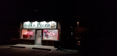 A small brick storefront is illuminated at night by overhead lights. The sign reads 'Bill's Pizza EST. 1957' with a pizza graphic, and there are lit neon signs in the window advertising 'Fabulous Hot Dogs' and 'Open Pizza'. The interior lights reveal some counters and equipment, and the area outside is dark with a few visible parking spots.