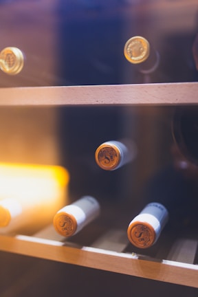 Several wine bottles are stored horizontally on a wooden wine rack within a glass cabinet. The corks and tops of the bottles are visible, and there is warm ambient lighting contributing to a cozy atmosphere.