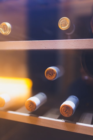 Several wine bottles are stored horizontally on a wooden wine rack within a glass cabinet. The corks and tops of the bottles are visible, and there is warm ambient lighting contributing to a cozy atmosphere.