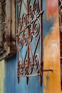 A detailed close-up of an ornate, rusted metal grill with intricate swirling patterns. The background contrasts the grill with a brightly-colored wall, blending shades of blue, orange, and yellow. The texture of the rust and the vibrant wall create a visually striking composition.