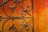 A close-up view of an ornate, wrought iron gate set against a vivid, textured orange-red wall. The ironwork features intricate swirling patterns, suggesting craftsmanship and age.