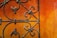 A close-up view of an ornate, wrought iron gate set against a vivid, textured orange-red wall. The ironwork features intricate swirling patterns, suggesting craftsmanship and age.