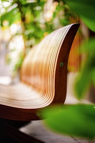 A close-up view of a modern, curved wooden bench with a smooth, polished finish. The bench is surrounded by lush green leaves, suggesting an outdoor or garden setting. The background is softly blurred, enhancing the focus on the bench's sleek design.
