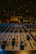 A close-up view of an audio mixing console with numerous faders and knobs, set in a dimly lit environment. Warm string lights softly illuminate the space, creating a cozy atmosphere. Blurred figures and a screen can be seen in the background, suggesting a live performance or event setting.