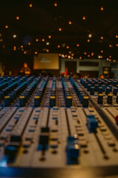 A close-up view of an audio mixing console with numerous faders and knobs, set in a dimly lit environment. Warm string lights softly illuminate the space, creating a cozy atmosphere. Blurred figures and a screen can be seen in the background, suggesting a live performance or event setting.