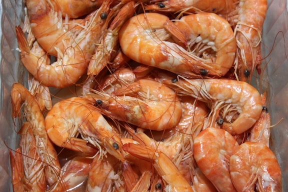 A close-up view of a pile of boiled shrimp with their shells still on, visible black eyes, and whiskers. The shrimp are laid out in a way that shows their pinkish-orange hue and shiny, slightly wet texture.